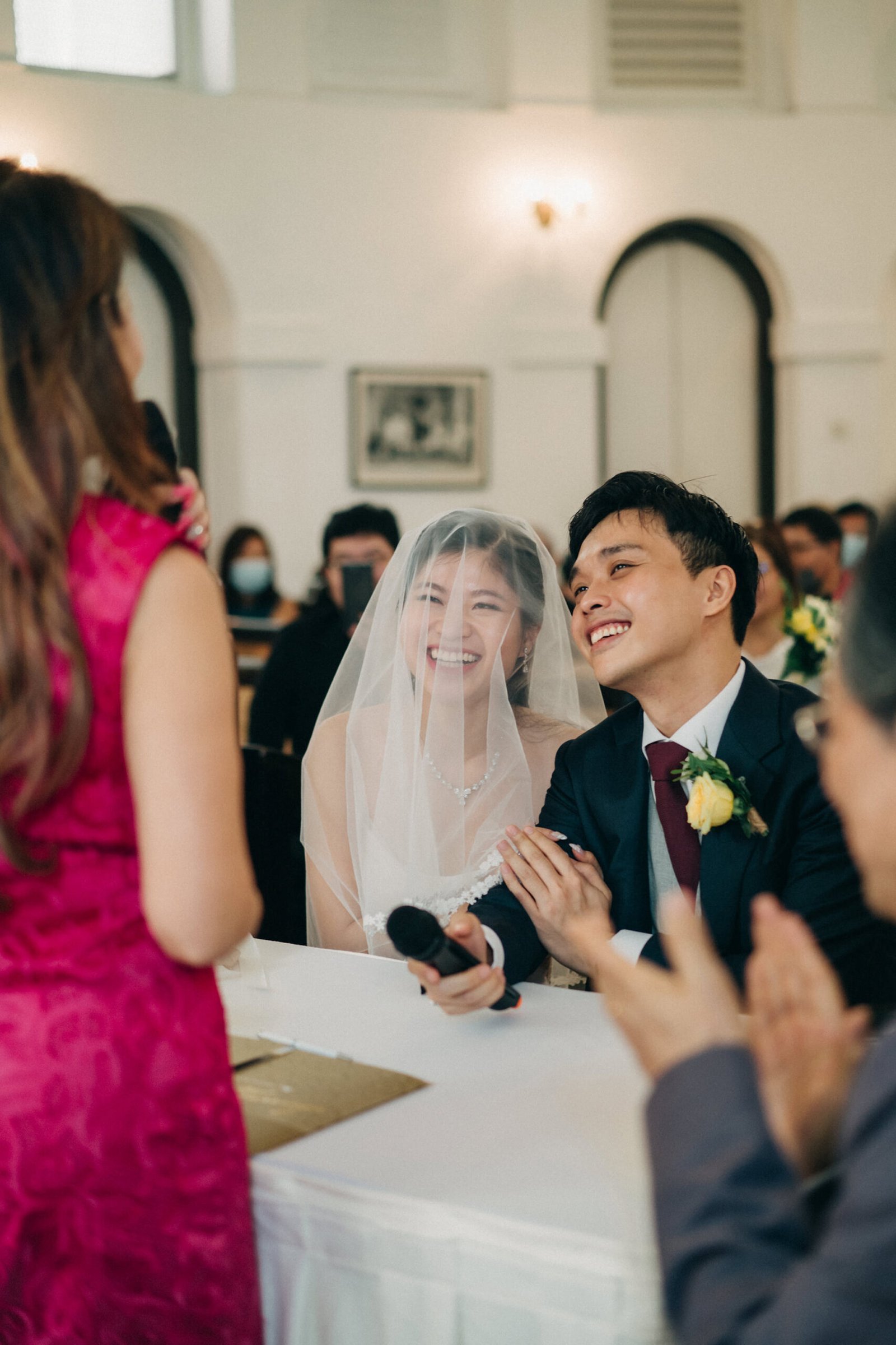 A joyous bride and groom laugh during their wedding ceremony, surrounded by guests. The bride wears a white veil, and the groom is in a dark suit with a red tie.