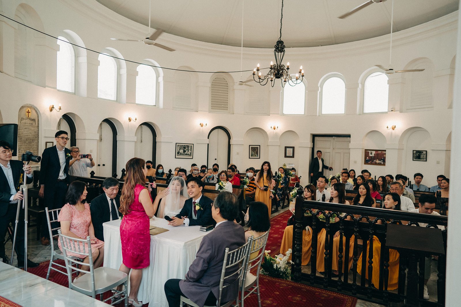 A wedding ceremony at Armenian Church Singapore unfolds in a bright, circular church with high arched windows. The bride in a white veil and groom sit at a table, surrounded by guests dressed in formal attire, with a vibrant pink dress standing out.