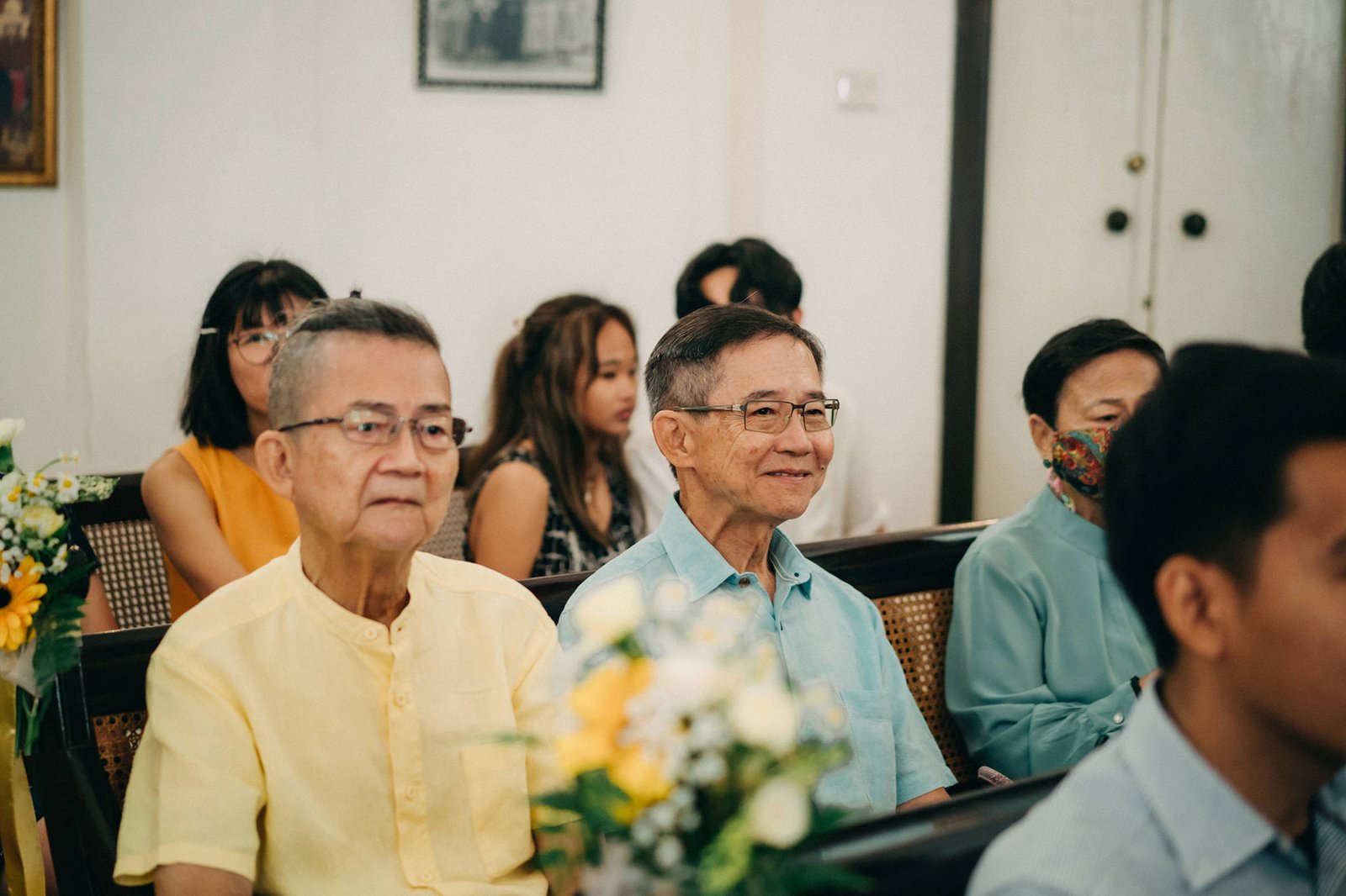 Elderly Asian men in light yellow and blue shirts, wearing glasses, smile during a gathering. Other guests, including women and younger men, are seated in the background, with floral arrangements adding a touch of color.