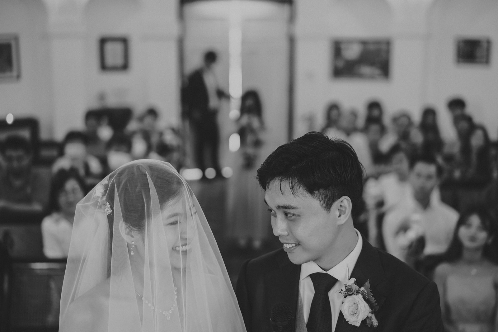 A joyous Asian couple, the bride in a veil and the groom in a suit, share a happy moment during their wedding ceremony, surrounded by guests in a church.