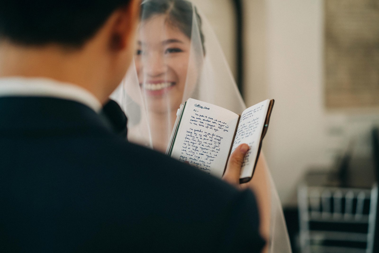 Bride in a veil smiles as she holds an open notebook with handwritten vows, facing her groom in a dark suit. The notebook has "Wedding Vows" written at the top.
