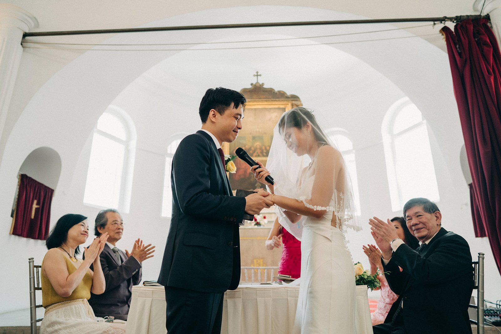 A bride and groom hold hands during a wedding ceremony, with the groom speaking into a microphone. Guests clap in the background, seated in a light-filled room with arched windows and red drapery.