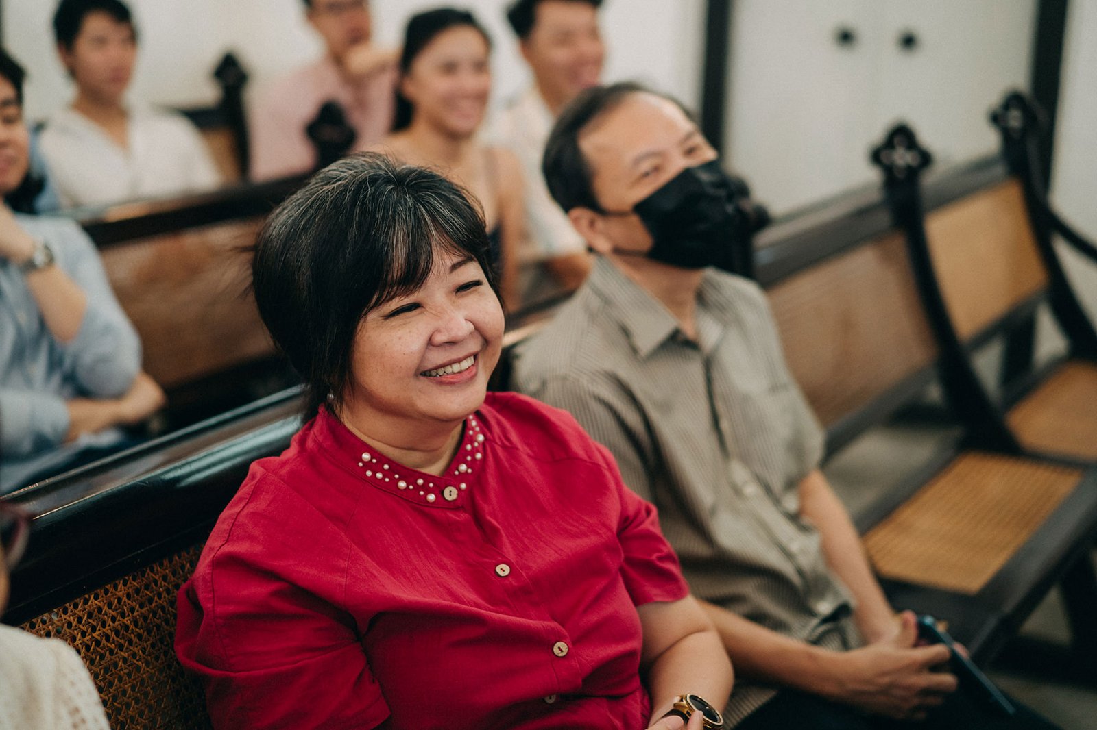 A smiling Asian woman in a red shirt sits in the front row of a church pew, looking to her left. Behind her, a man in a striped shirt and black mask laughs.