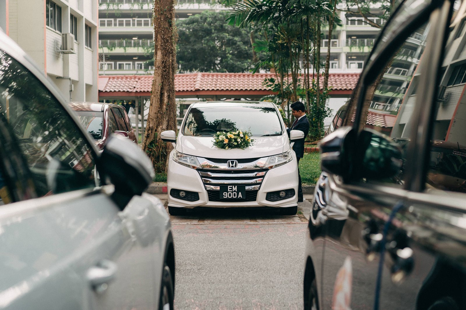 A white wedding car, adorned with a floral arrangement on its hood, is parked between two other cars. The license plate reads "EM 900 A". A man in a suit stands near the car.