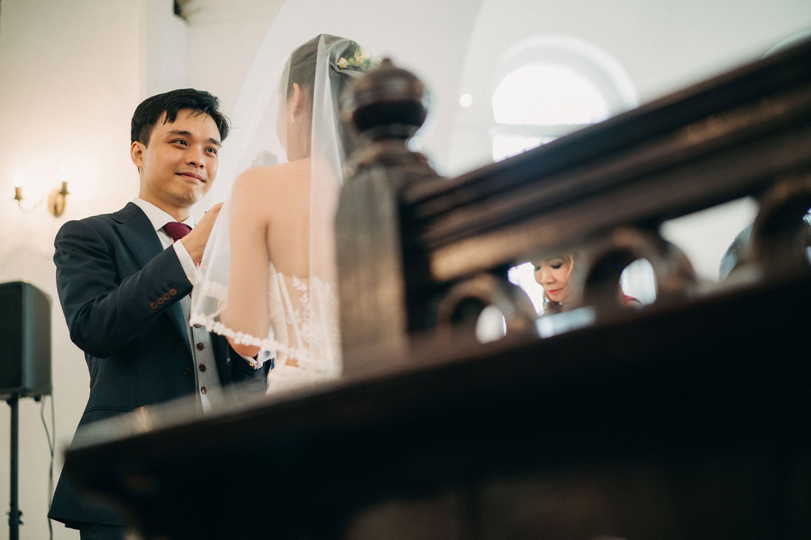 Groom in a dark suit and burgundy tie adjusts his bride's veil, both smiling tenderly during their wedding ceremony in a dimly lit church with arched windows.