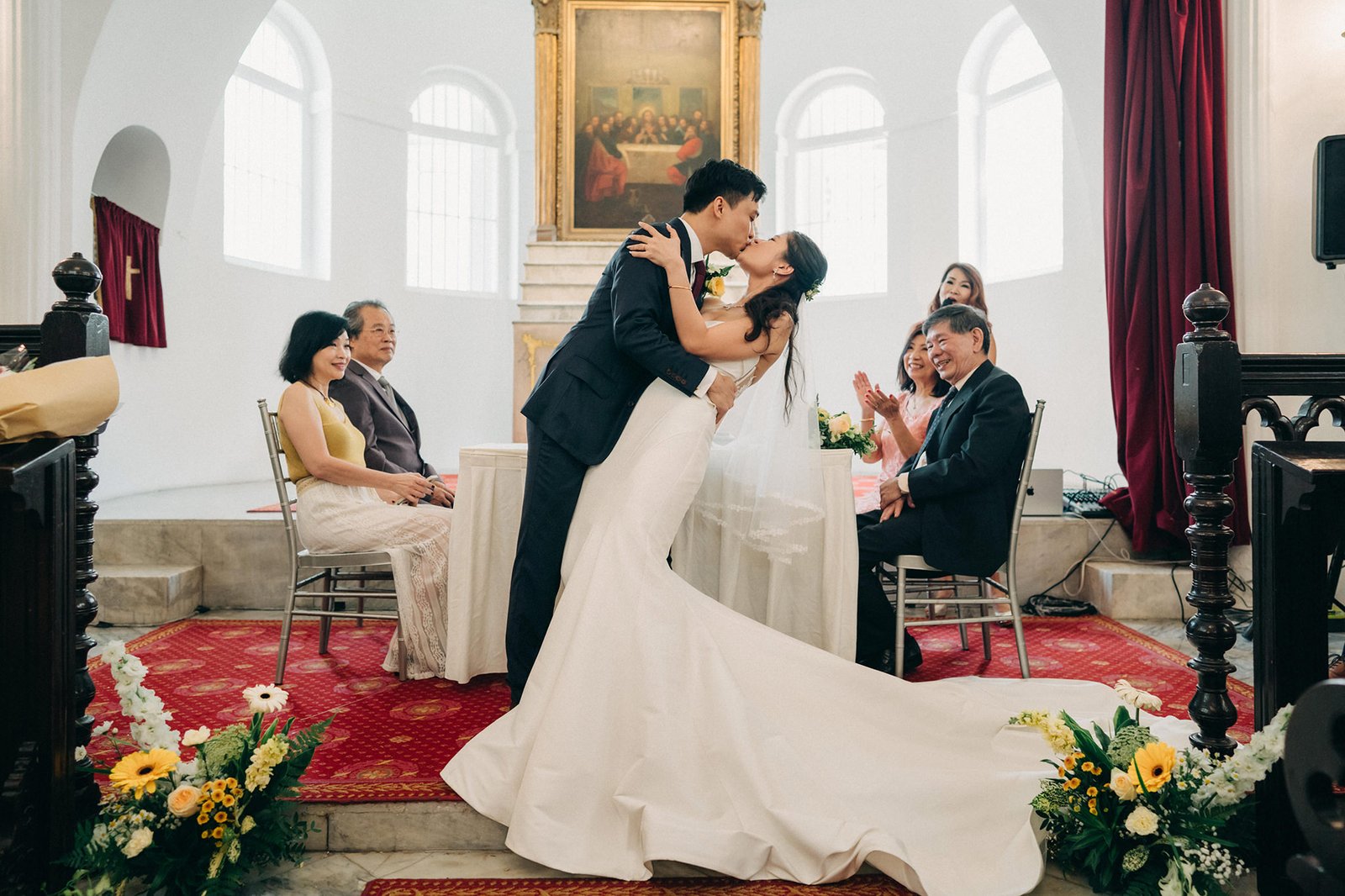 A bride in a stunning white mermaid wedding dress kisses her groom in a church ceremony, surrounded by family and floral arrangements.