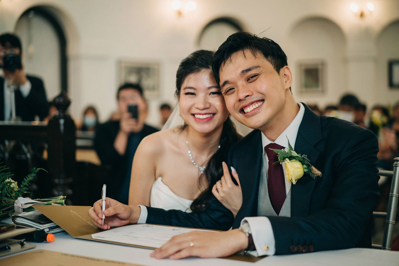A smiling Asian couple, a bride in a white strapless dress and a groom in a dark suit and vest, embrace at a wedding ceremony table, signing documents. The groom has a yellow boutonniere.