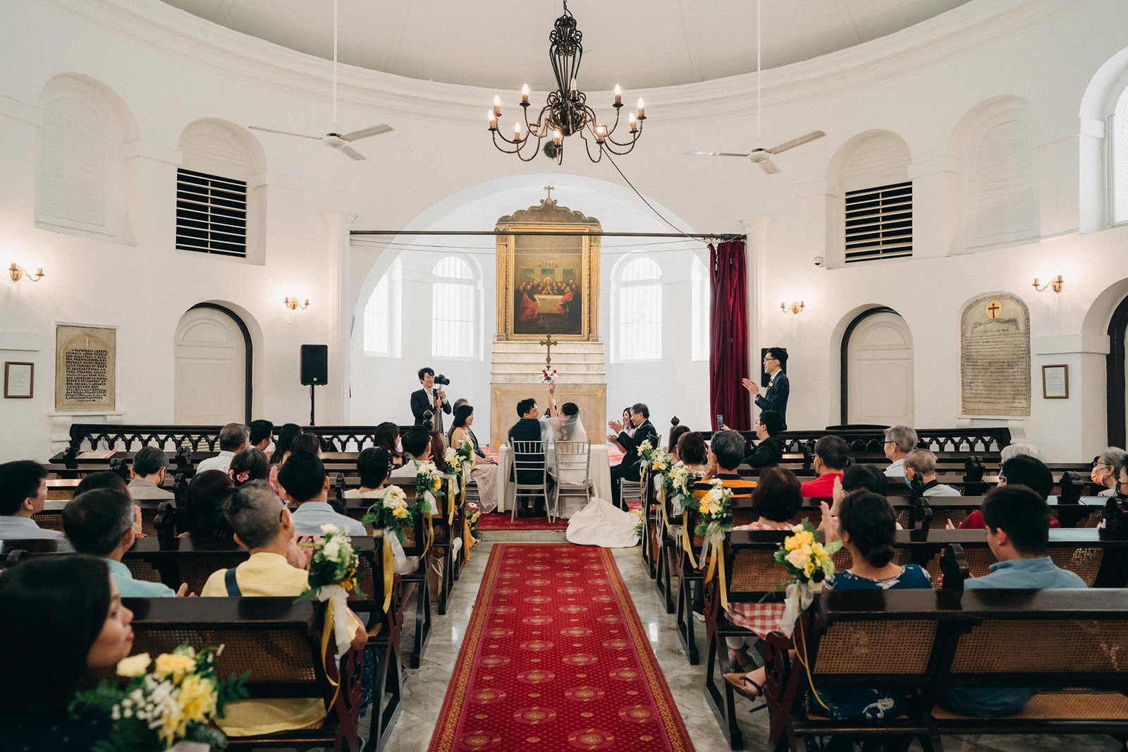 A wedding ceremony in Armenian Church Singapore with red carpet aisle, bride and groom at altar, guests seated in pews, and a painting above the altar.