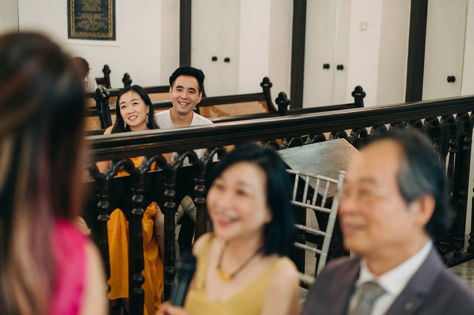 A smiling Asian couple sits in the foreground of a wedding ceremony, with blurred guests in front and behind them. The bride wears a yellow dress, and the groom wears a white t-shirt.