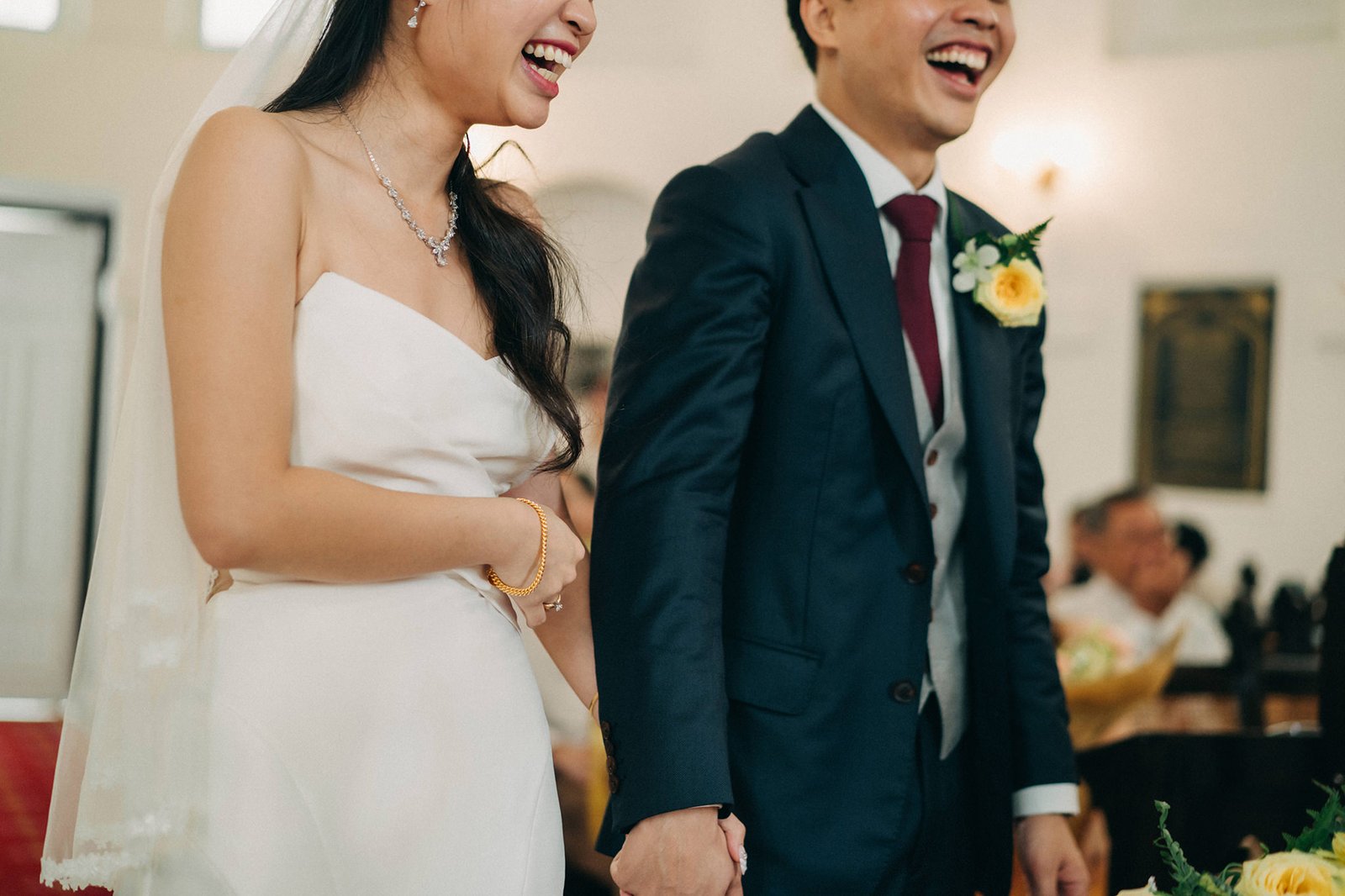 A joyful bride in a white strapless gown and a groom in a dark suit laugh heartily, holding hands during their wedding ceremony. A yellow rose boutonniere adorns the groom's lapel.