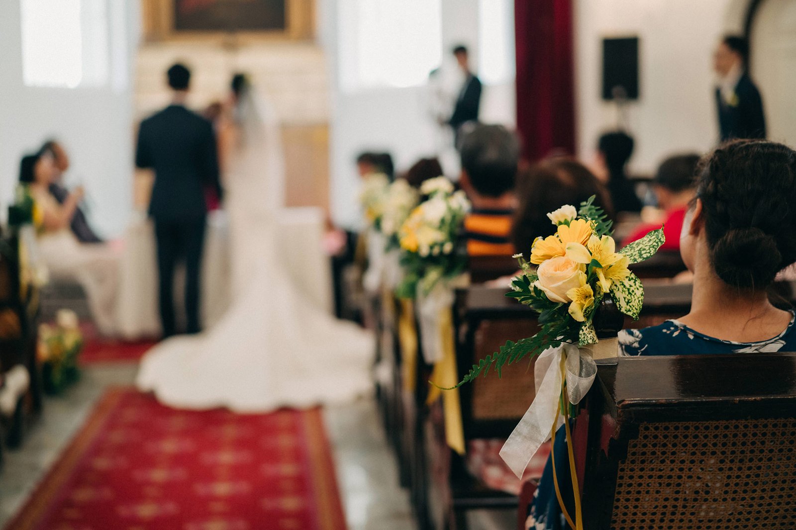 A wedding ceremony at Armenian Church Singapore with guests seated in pews. A bridal bouquet of yellow and white roses is tied to a pew in the foreground, with the bride and groom blurred in the background.