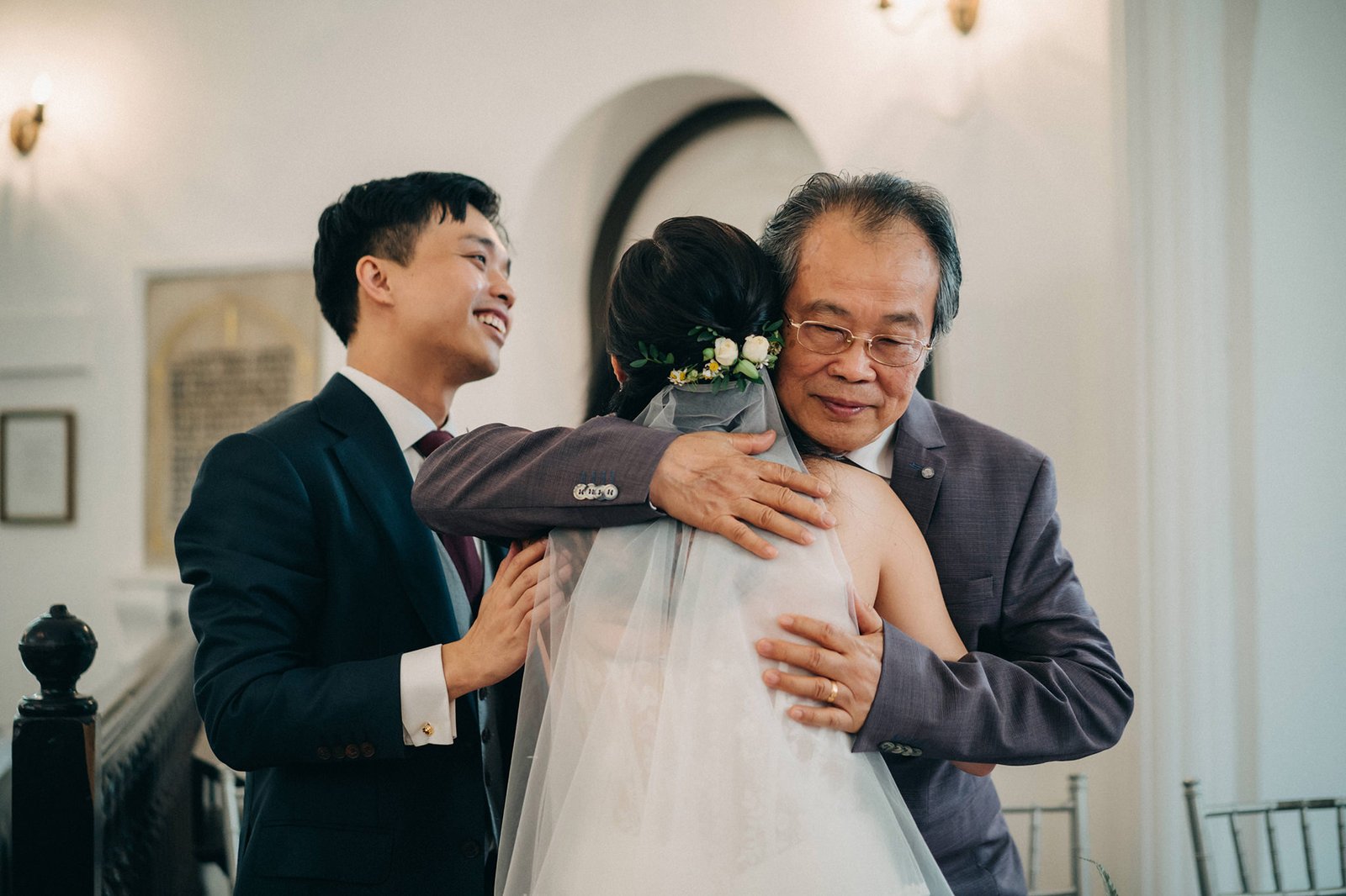 A proud father in a grey suit embraces his daughter, the bride in a white wedding dress, during her wedding ceremony. The groom, in a dark suit, smiles joyfully in the background.