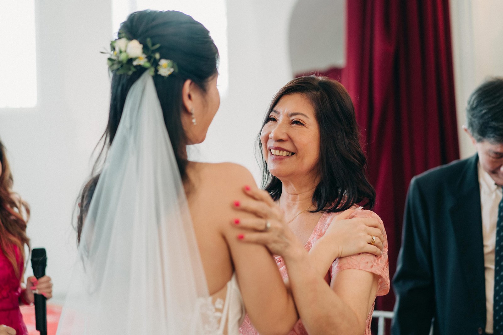 A smiling mother in a pink dress embraces her daughter, the bride, who wears a white wedding gown and veil adorned with flowers. The emotional moment captures a wedding celebration.