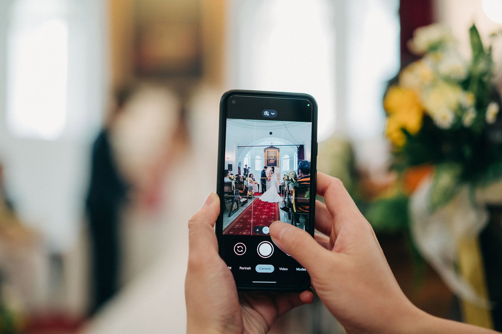 A person's hands hold a smartphone, capturing a wedding ceremony in a church with a red carpet and floral decorations. The phone screen displays the live camera view of the bride and groom.