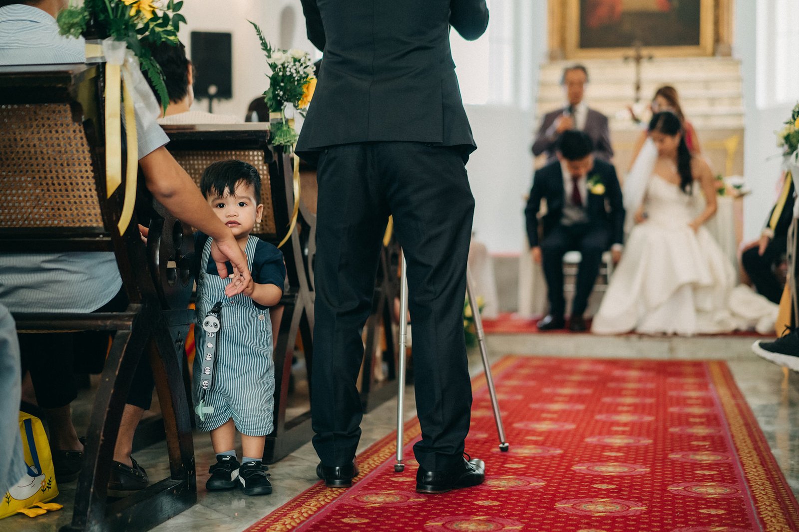 A young boy in blue overalls stands on a red carpet at a wedding ceremony, holding a small umbrella. In the background, a bride and groom are visible with guests seated in the pews.