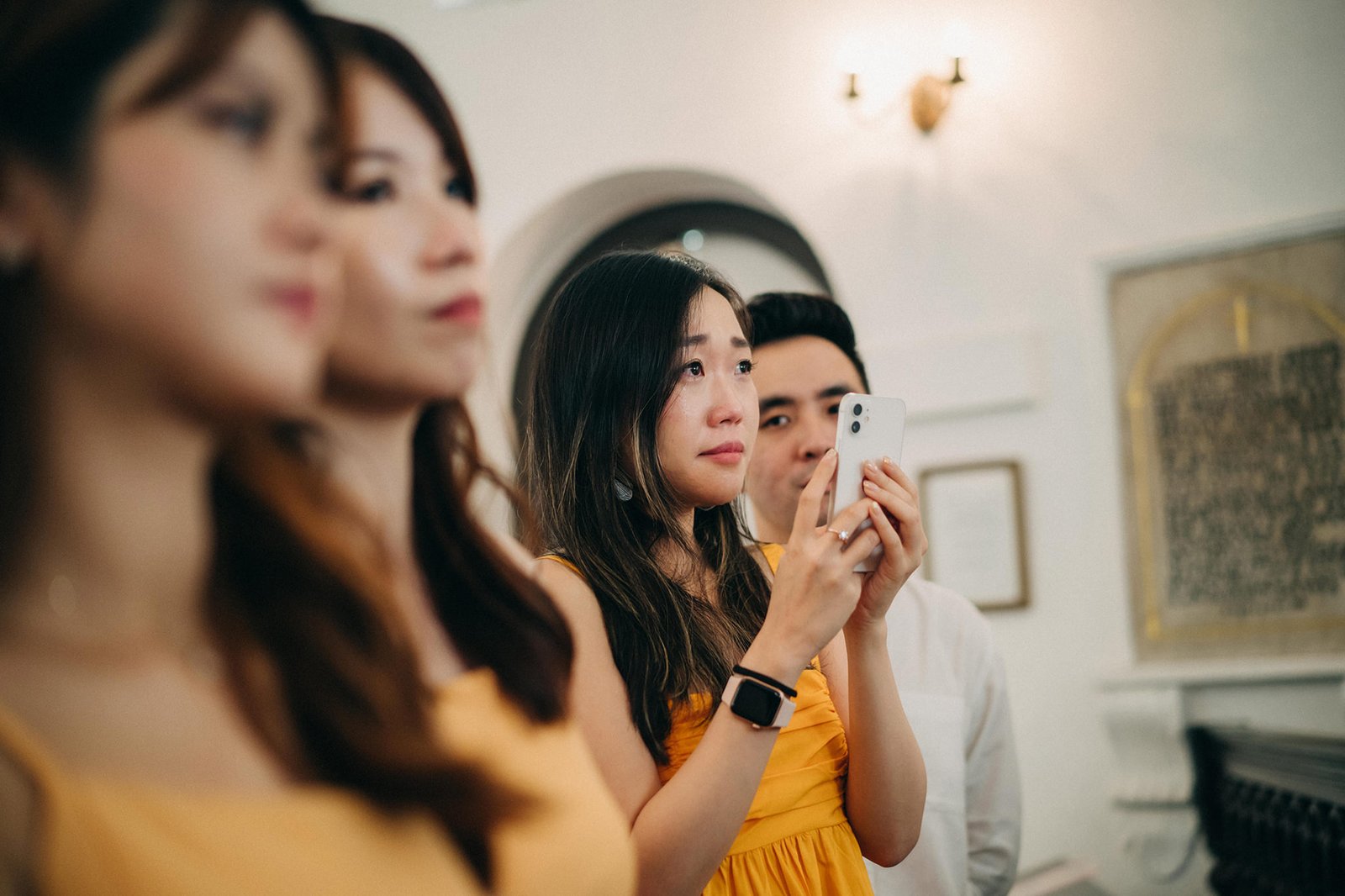 A tearful young woman in a yellow dress records a wedding ceremony on her smartphone, surrounded by other wedding guests.