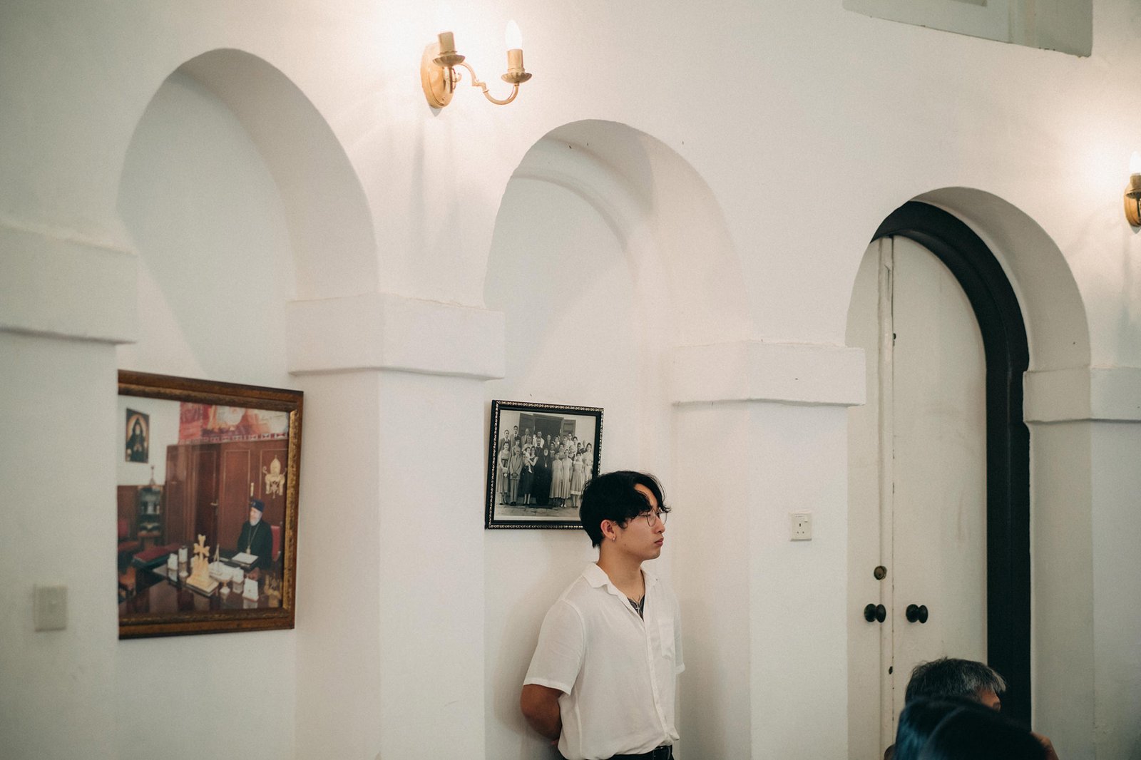Young man in white shirt stands in a white arched church interior, looking to the side, with framed historical photos on the wall.