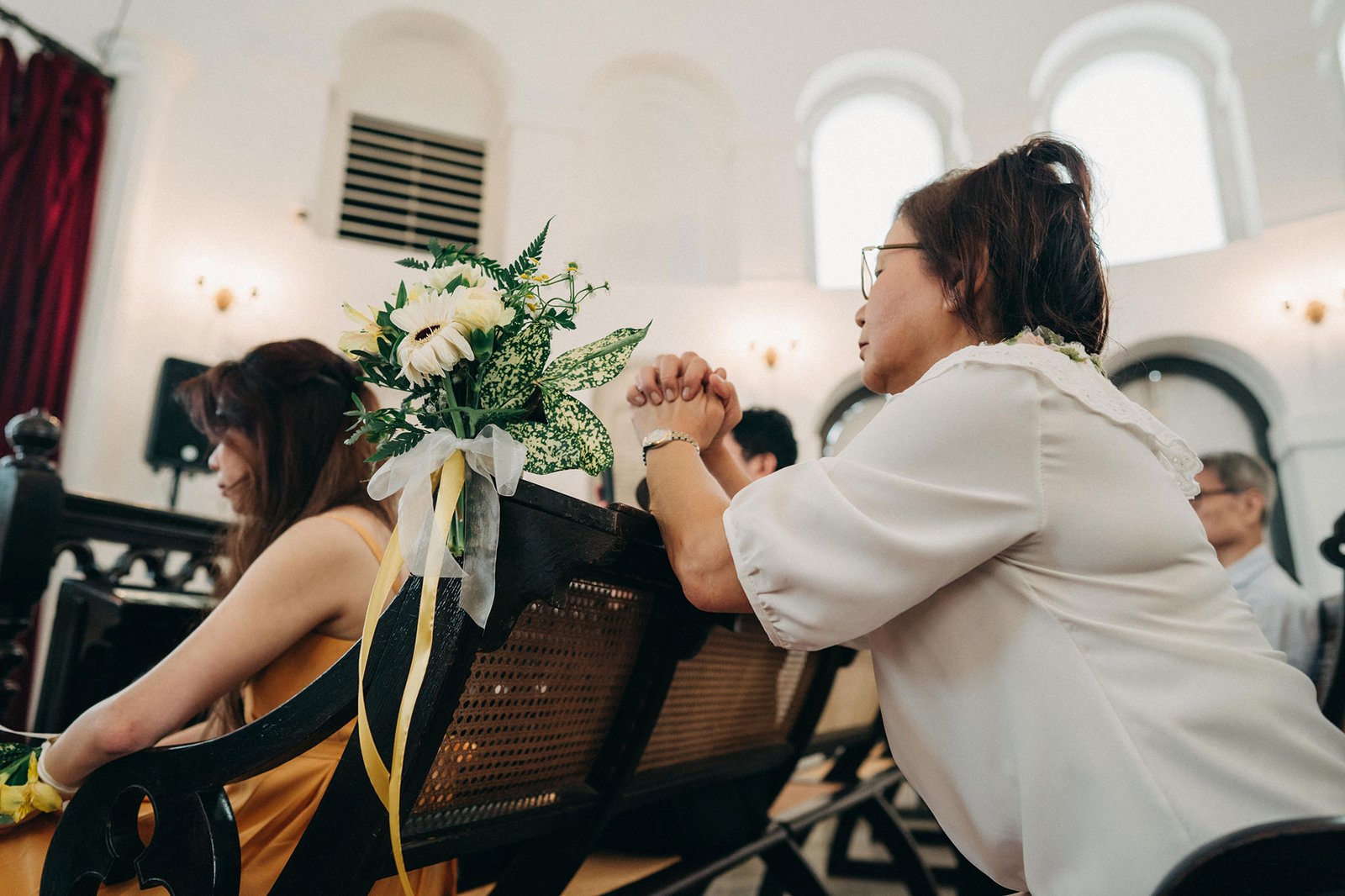 Two women attend a wedding ceremony, one in a yellow dress and the other in a white top, with a floral arrangement on a pew in the foreground.