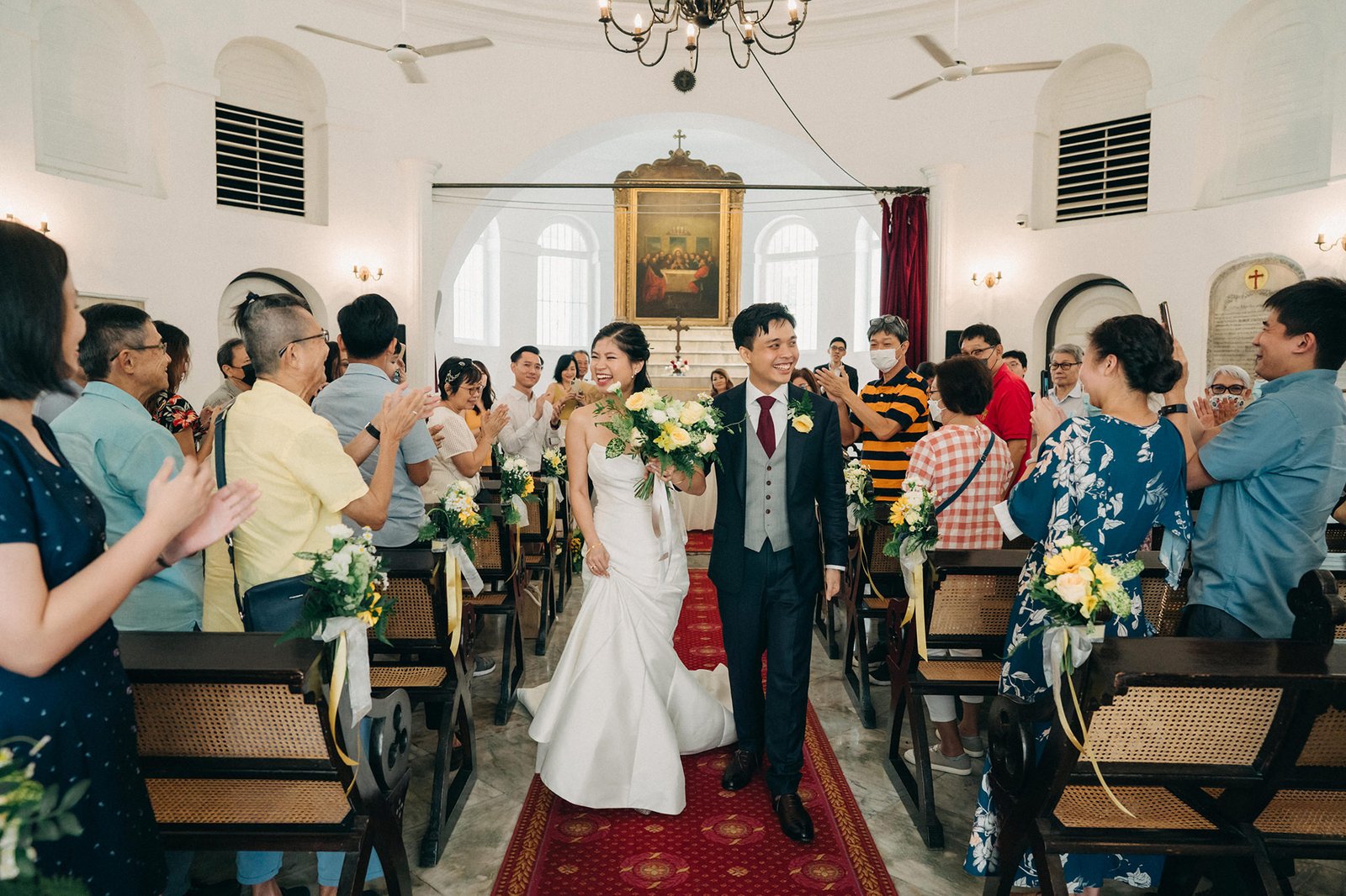 Newlyweds walk down the aisle of a church at Armenian Church Singapore, smiling and waving to guests. The bride wears a white wedding dress, and the groom is in a dark suit. Guests are seated on wooden benches, and yellow flowers adorn the aisle.
