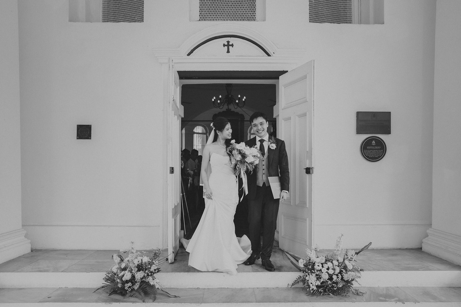 A smiling bride in a strapless gown and a groom in a suit and tie exit a church, holding a bouquet, with floral arrangements flanking the doorway.