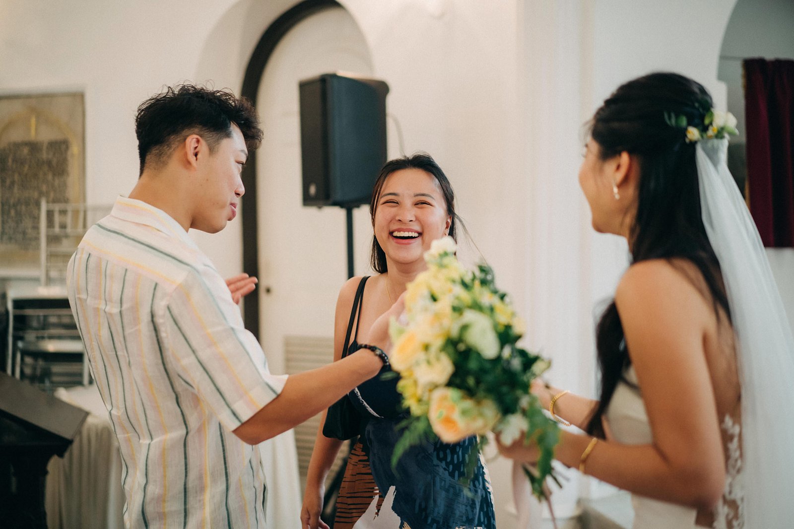 A bride in a veil receives a bouquet from a smiling couple.