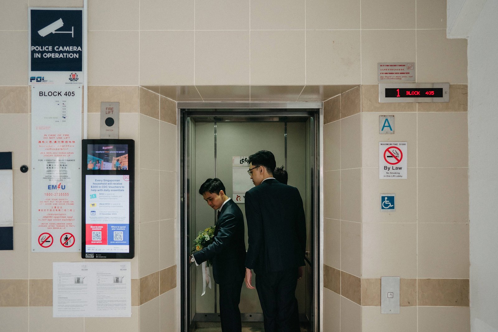 Elevator doors open to reveal two men in dark suits, one holding a bouquet of flowers, entering an elevator in a building with "BLOCK 405" signage.