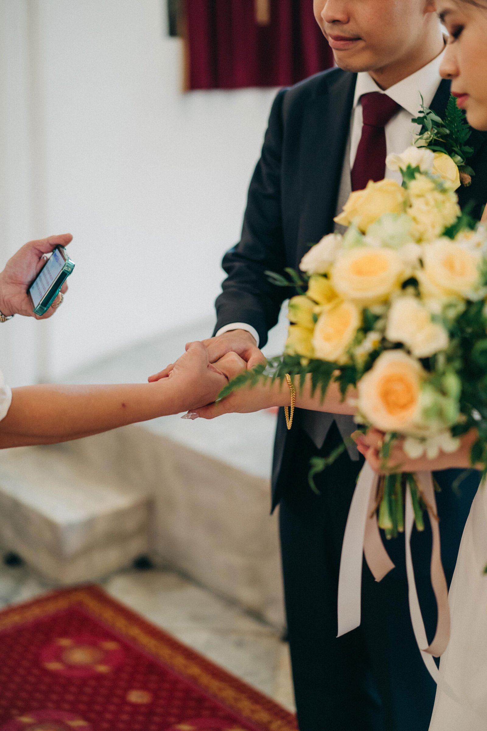 A wedding ceremony: a couple holds hands, a bouquet of yellow and white roses is visible, and someone is holding a phone to record the event.