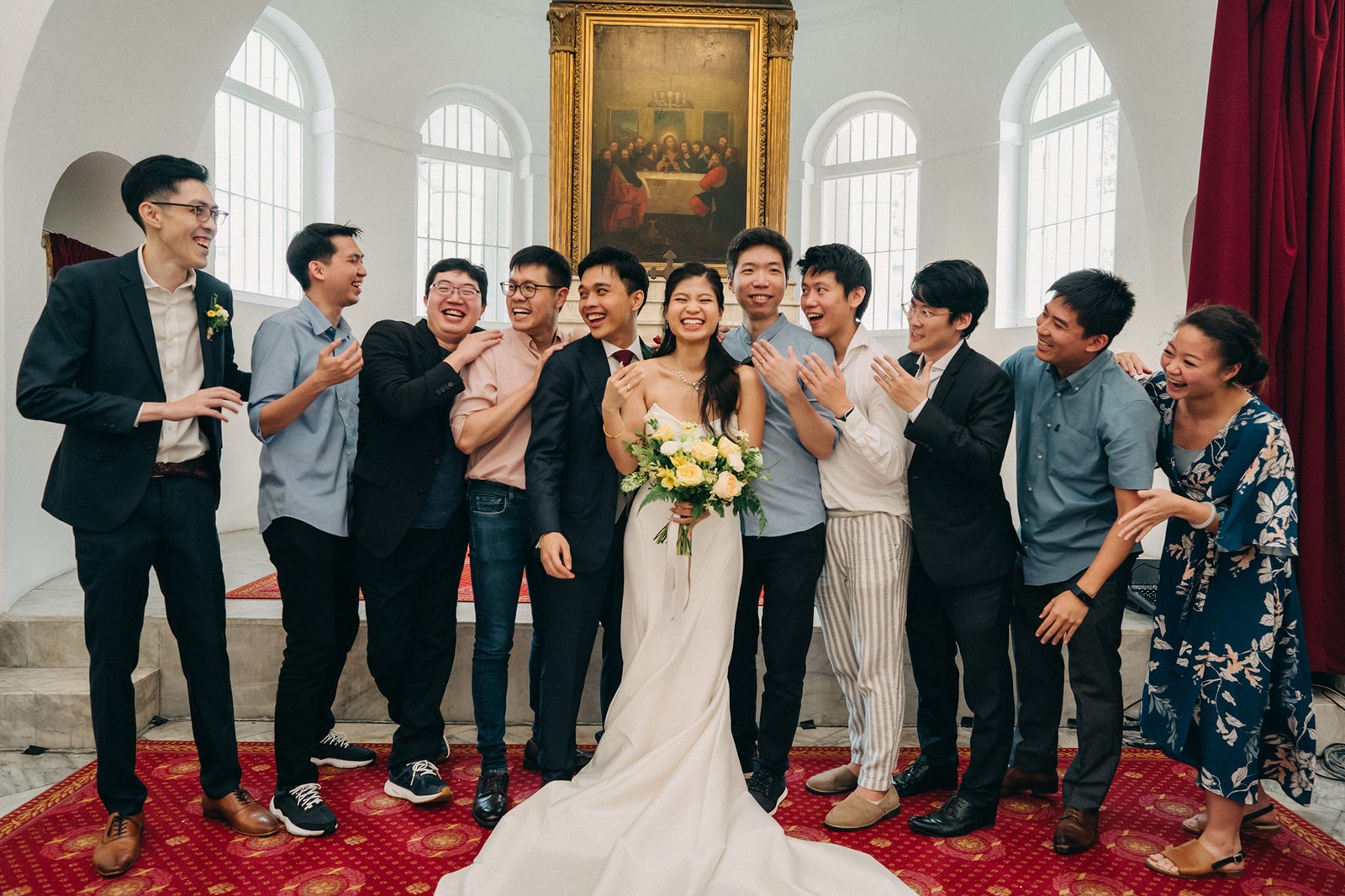 A diverse wedding party poses for a photo in a grand room with tall windows, a large painting, and red velvet curtains. The bride and groom are in the center, surrounded by smiling groomsmen and bridesmaids in casual attire.