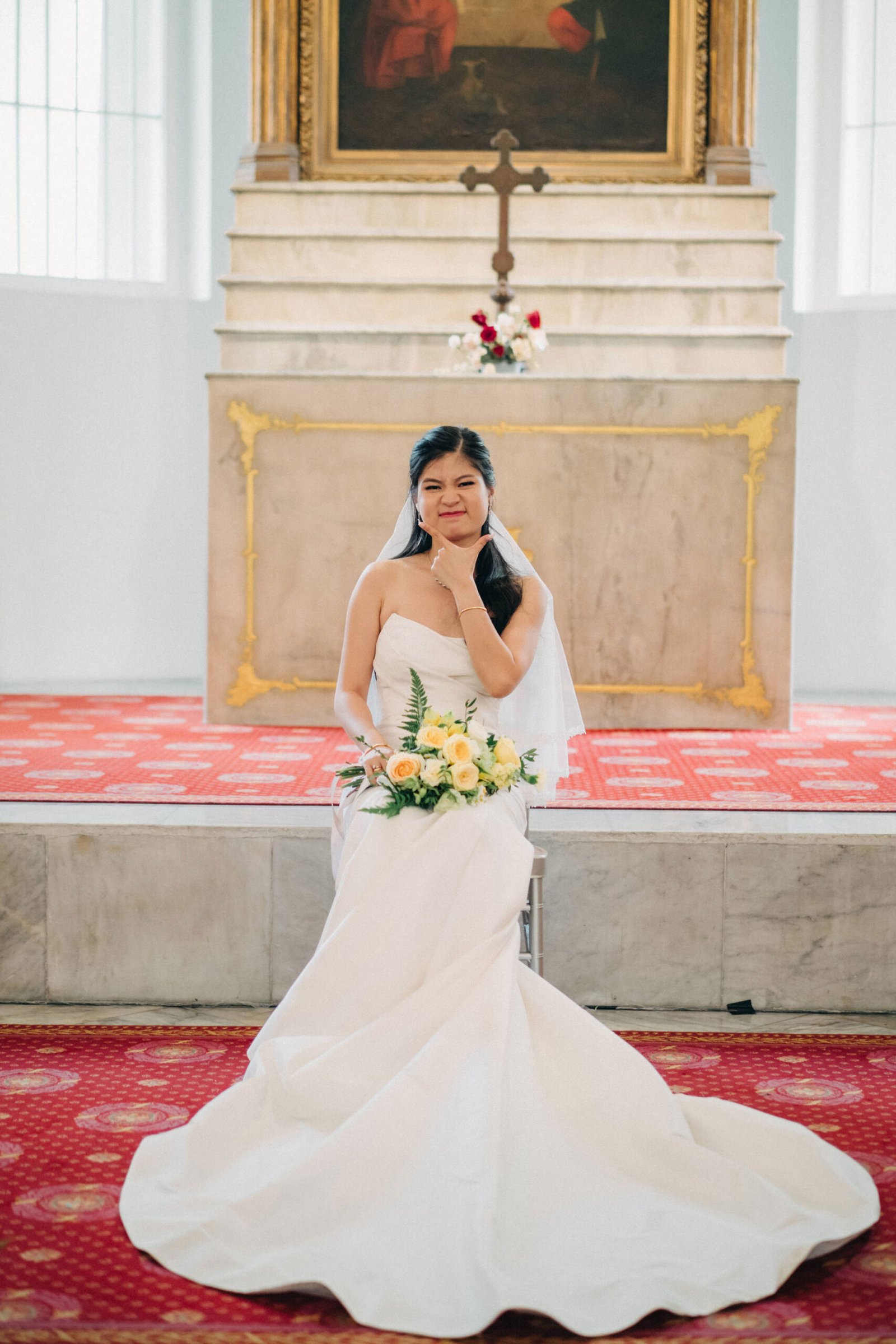 A bride in a strapless white wedding dress kneels with a bouquet of yellow and white roses in a church, with a gold-framed altar and red carpet visible.