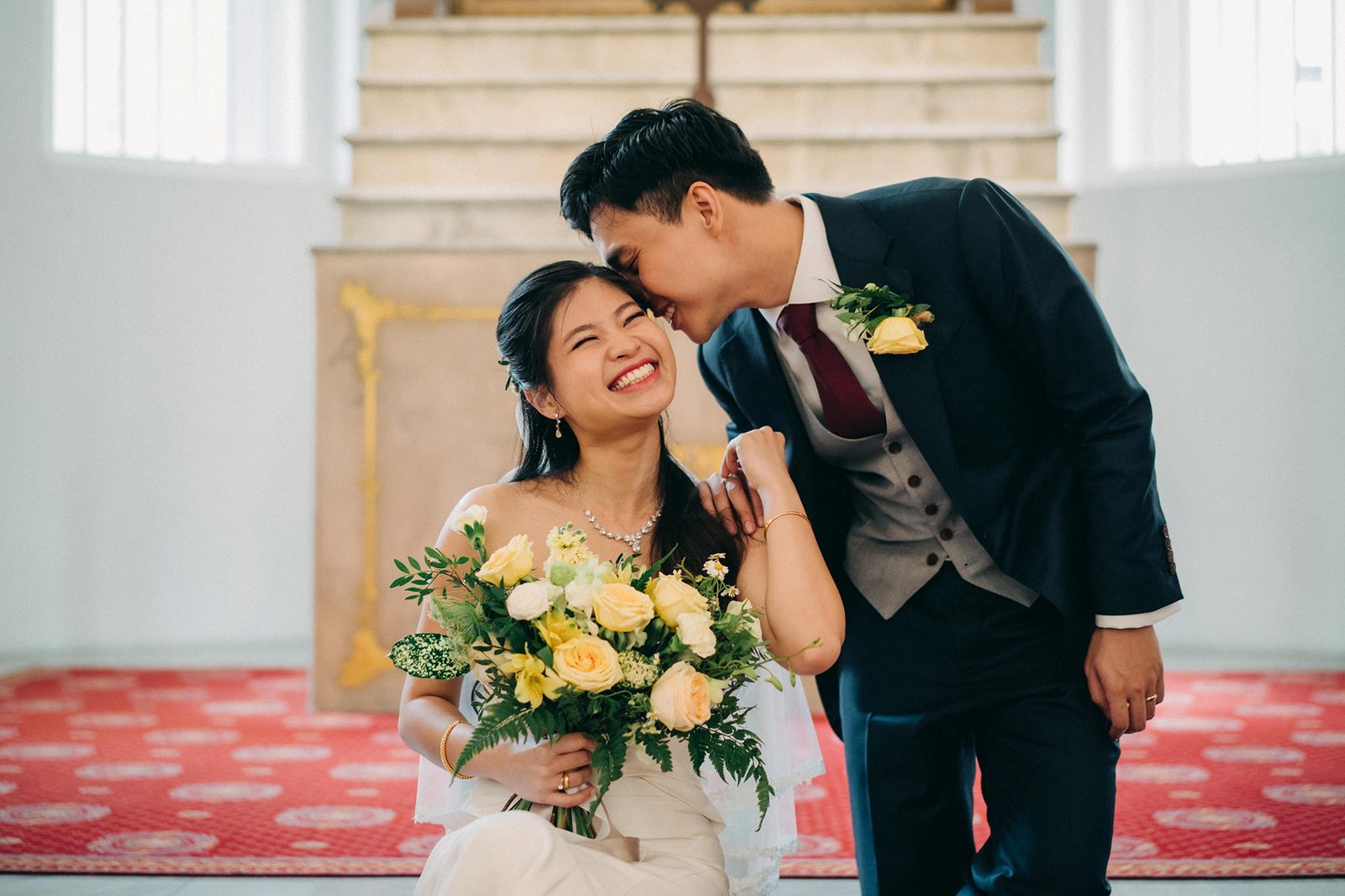 A joyful Asian bride in a white gown laughs as her groom in a suit whispers in her ear, holding a bouquet of yellow and white roses. The wedding ceremony takes place in a church with red carpet and ornate stairs.