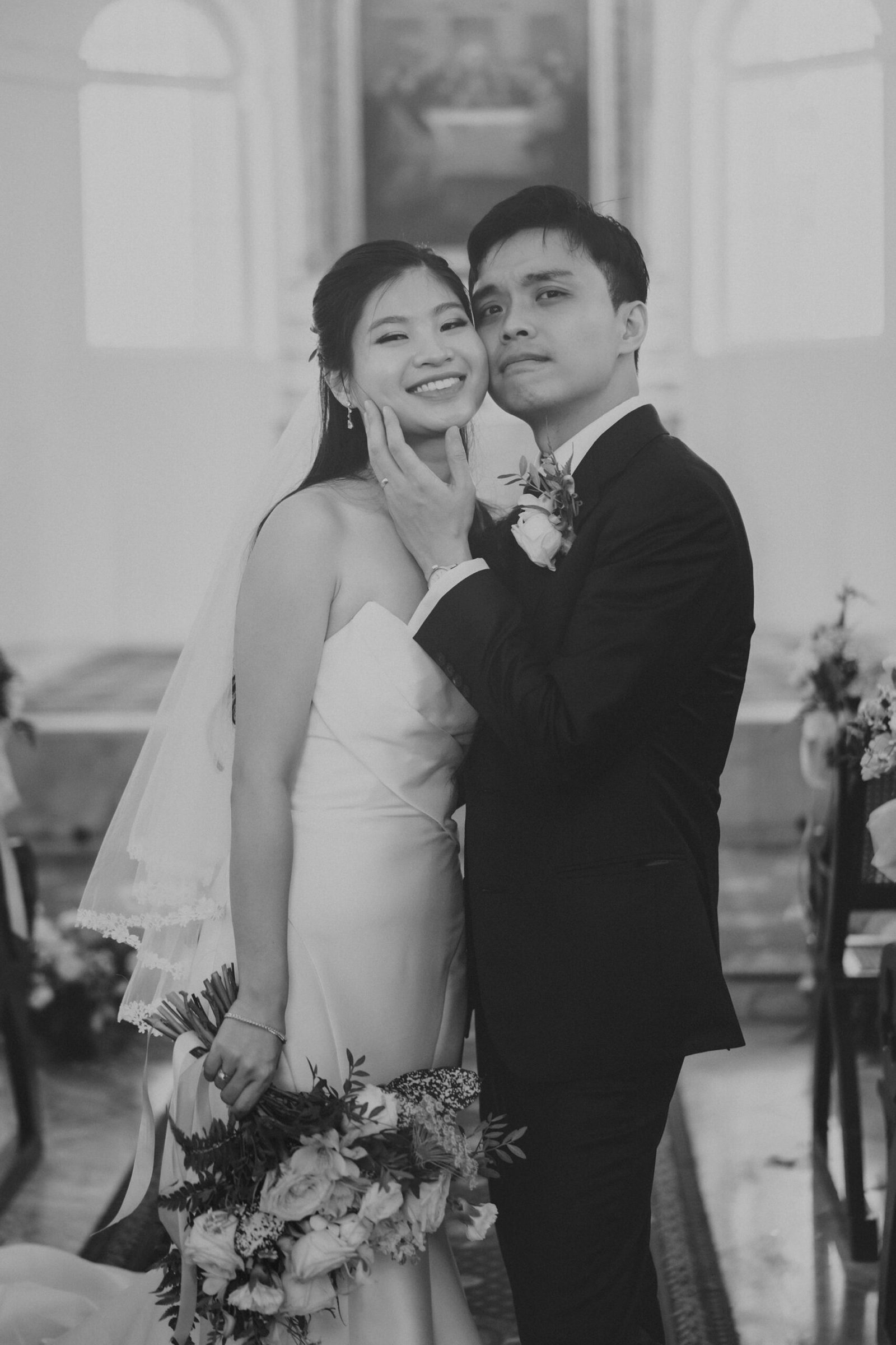 A joyful bride in a strapless gown and a groom in a tuxedo share a tender moment, embracing in a church aisle adorned with flowers. The black and white photograph captures their wedding day smiles at Armenian Church Singapore.