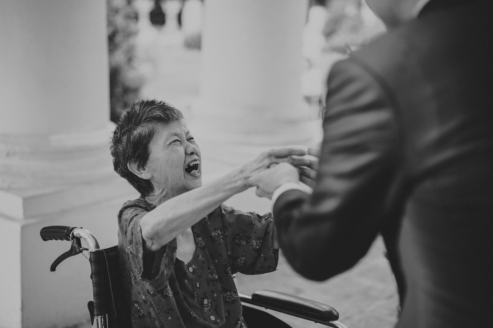 Elderly woman in wheelchair laughing and reaching out to a man in a suit, capturing a moment of joy and connection.