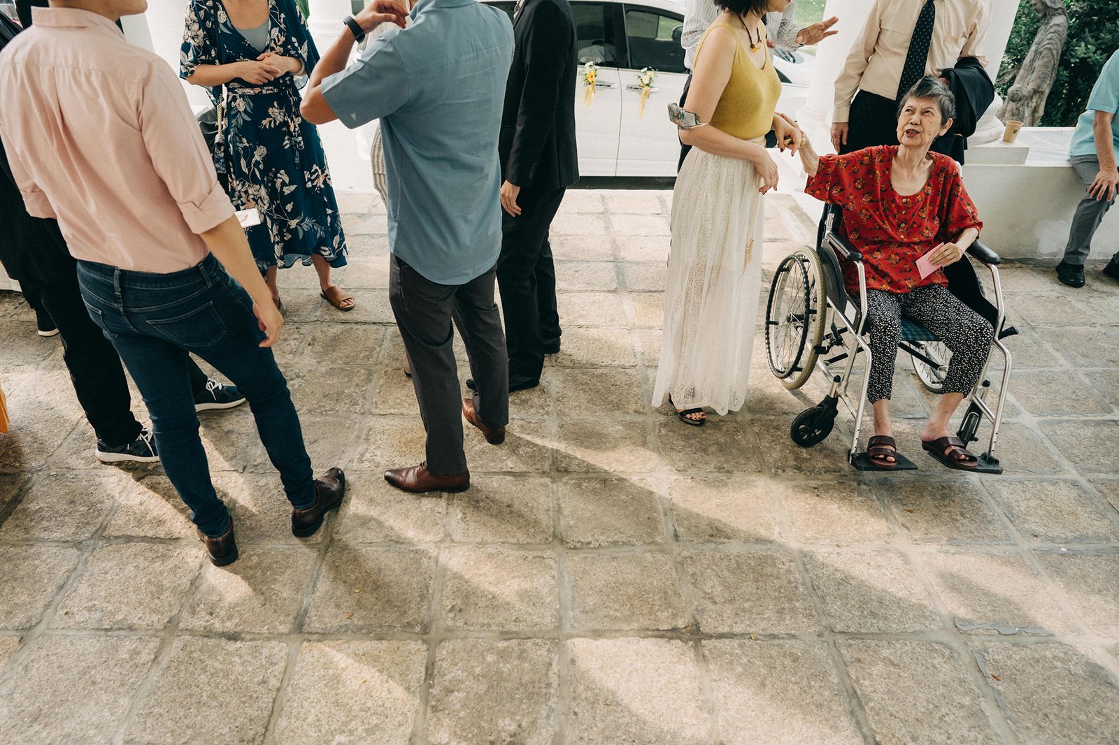 A diverse group of people, including an elderly woman in a wheelchair, gather outdoors on a stone patio. The woman wears a red top and is being attended to by others in casual attire. The scene suggests a social gathering or event.