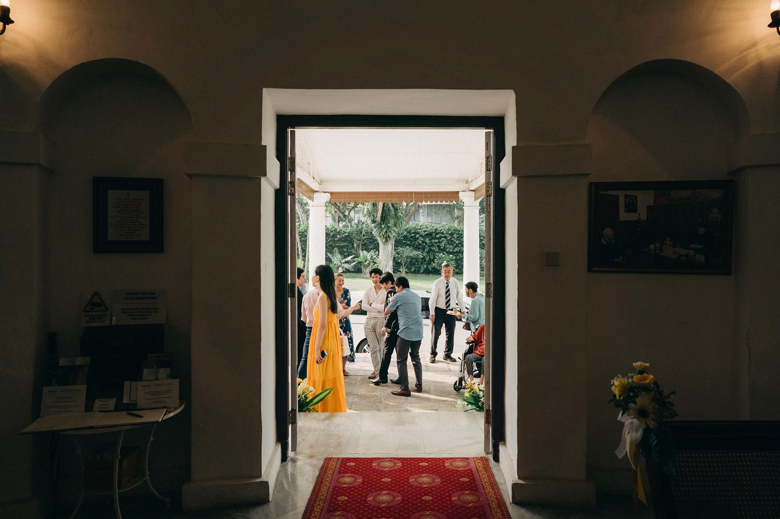 A wedding party gathers in a sunlit courtyard through an arched doorway, with guests in formal attire mingling near a woman in a vibrant yellow dress.