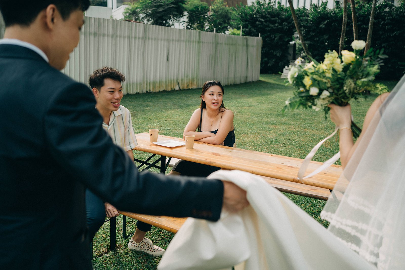 A wedding couple and guests are gathered outdoors at a wooden picnic table, with the bride holding a bouquet and veil. The scene is set on a grassy lawn with a white fence in the background.