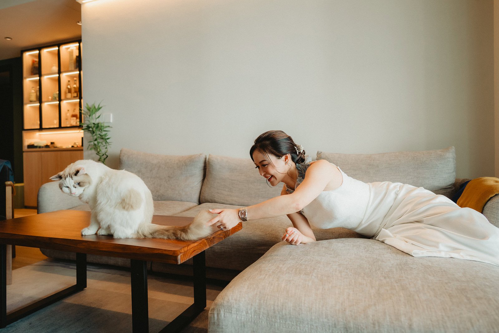 Bride in white dress plays with fluffy cat on sofa at Capella Singapore wedding